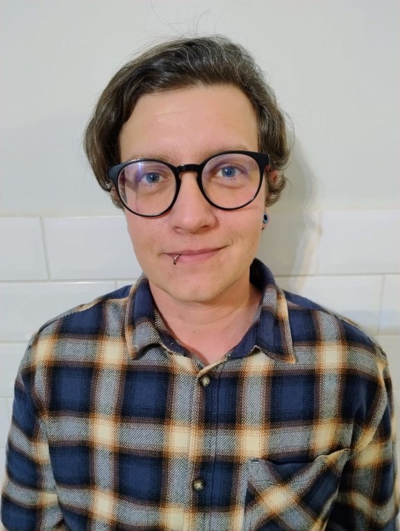 A white person with short, side-parted brown hair, blue eyes and wear round glasses with a black frame, red sides, and a gray piercing on the right corner of their mouth. They are wearing a dark blue and white plaid shirt.