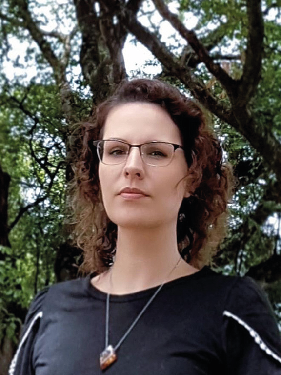 Caucasian woman, light brown hair, wavy hair, shoulder length, wearing black framed glasses, using a black t-shirt with a pendant necklace and trees in the background