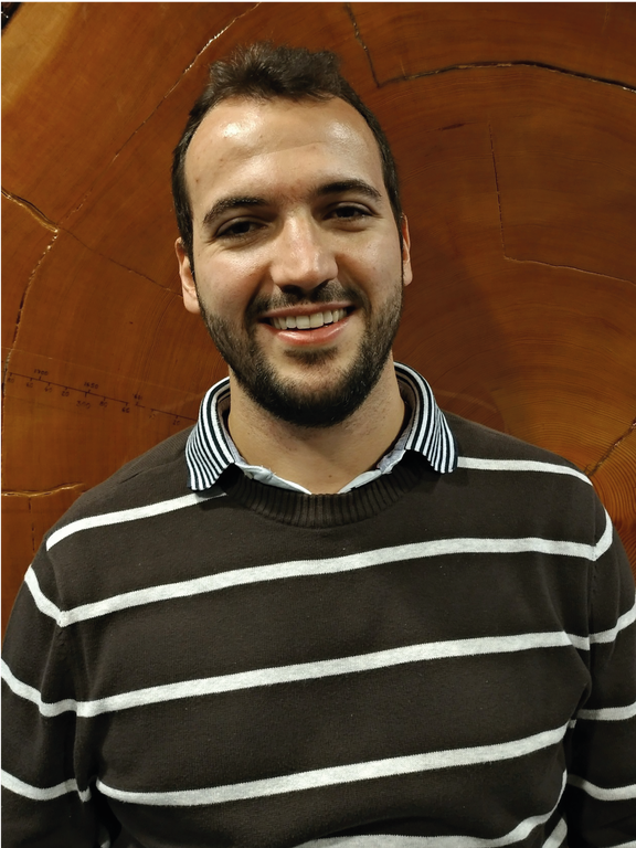 White, short brown haired, unshaven faced male smiling, wearing a white-striped brown shirt, and standing in front of the cross-section of a Douglas fir trunk