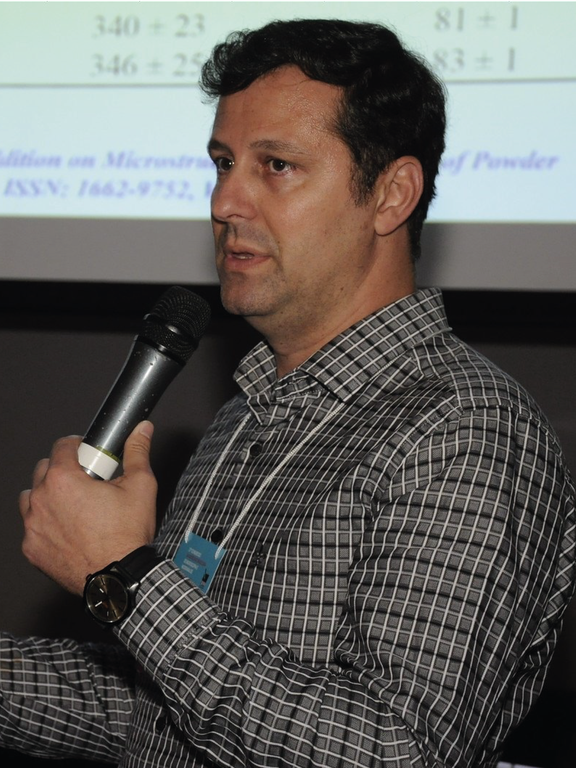 Man, white, brown eyes, brown hair, short and straight, wearing a black, gray and white shirt and at microphone