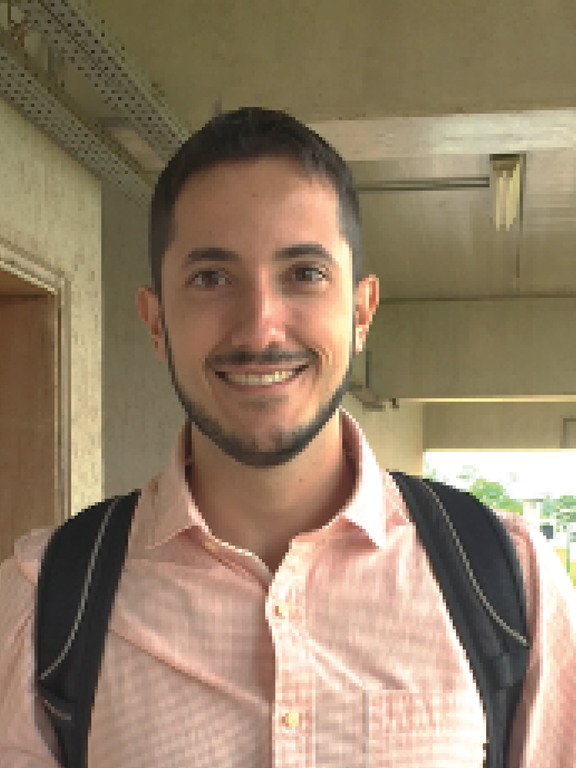 Man, white, with short dark hair at the sides and a little longer at the top, a thin beard around his chin and goatee, a beard shaved on his cheeks and neck. Smile for having just been hired as professor at UFSCar, wearing a pink shirt with only the collar open, a little flushed due to the hot day, black backpack straps on the shoulders. Although the photo shows only the face, you can see the corridor in the background, it is the corridor of the AT6 of UFSCar, the day is sunny.