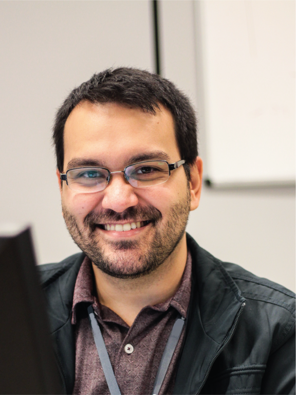 Man, white, short black hair, beard, wearing rectangular black glasses, with a black jacket and a red polo shirt