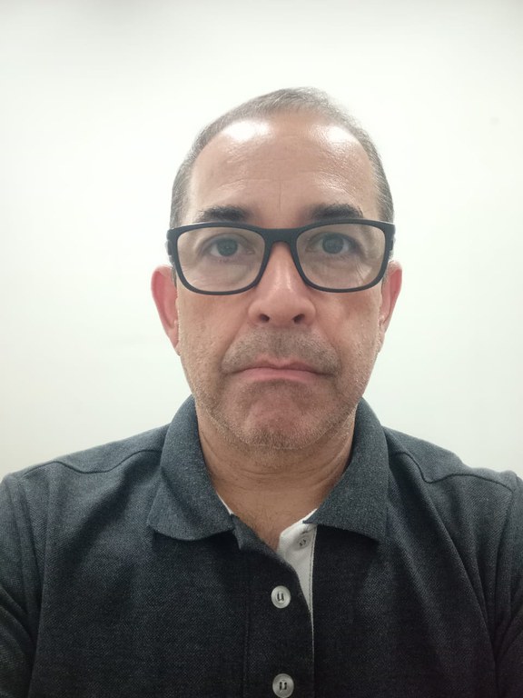 Homem branco, cabelos curtos e grisalhos. Ele usa óculos de grau com armação grossa preta e retangular. Veste camisa polo de cor cinza escuro mescla, com um detalhe branco visível na parte interna do colarinho e botões claros.