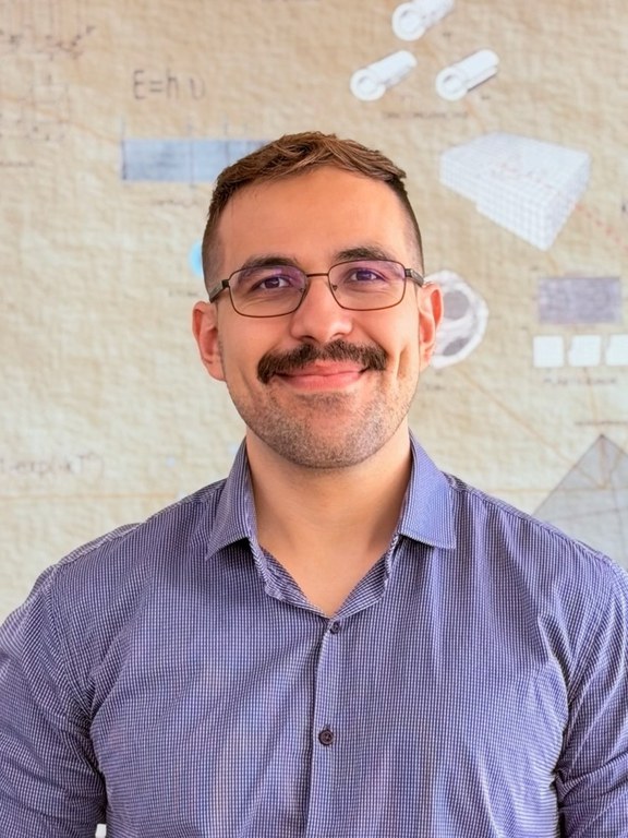 Homem, branco, cabelo castanho liso e curto, bigode preto, usando óculos com armação preta, com camisa azul quadriculada. Ao fundo, painel do saguão do DEMa - UFSCar, apresentando o tetraedro da engenharia de materiais, discordâncias e a equação de Planck. 